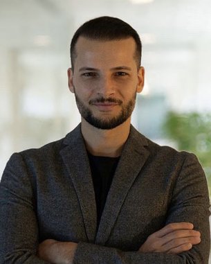 Ein Mann mit kurzem Bart und kurzen Haaren steht mit verschränkten Armen in einem modernen Raum, umgeben von Pflanzen. Er trägt einen grauen Blazer und blickt direkt in die Kamera.