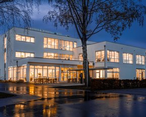 Moderne, mehrstöckige Gebäude mit großen Fenstern, beleuchtet bei Dämmerung. Nasser Parkplatz im Vordergrund spiegelt die Lichter wider.