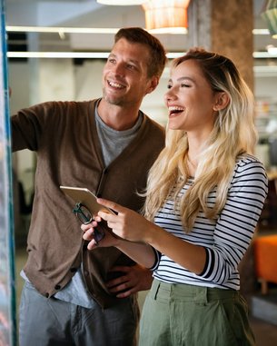 Zwei Personen stehen vor einem interaktiven Bildschirm, auf dem Diagramme und Notizen zu sehen sind. Die Frau hält ein Tablet und lächelt, während der Mann interessiert auf den Bildschirm schaut.