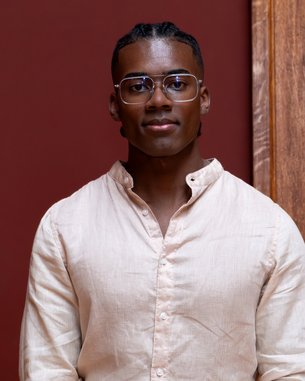 Ein junger Mann mit kurzen, geflochtenen Haaren und Brille trägt ein helles Hemd und steht vor einer roten Wand.