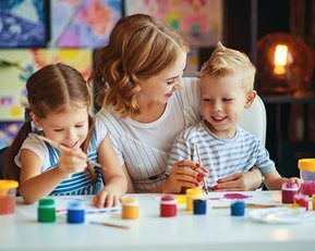 Eine Frau sitzt mit zwei Kindern an einem Tisch, während sie gemeinsam malen. Die Kinder verwenden Pinsel und Farben, um Bilder zu gestalten. Der Tisch ist mit verschiedenen Malutensilien und Farben bedeckt.