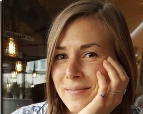 Eine Person mit kurzen, braunen Haaren sitzt an einem Tisch und stützt ihr Gesicht mit der Hand. Sie trägt ein gemustertes Hemd.