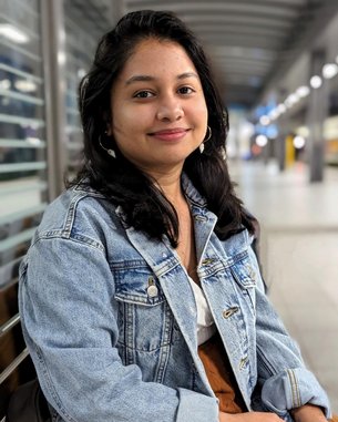 Eine junge Frau mit langen, dunklen Haaren sitzt auf einer Bank, trägt eine Jeansjacke und lächelt in einem modernen Innenraum.