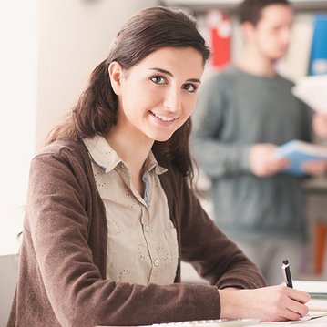 Fernstudium Bachelor Soziale Arbeit | SRH Fernhochschule Eine junge Frau sitzt an einem Tisch und schreibt, während zwei Personen im Hintergrund stehen und Unterlagen halten. Der Tisch ist mit Schreibutensilien und einem Notizbuch ausgestattet.