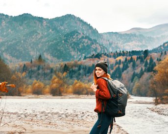 Eine Person mit einem Rucksack steht am Ufer eines Flusses, umgeben von herbstlichen Bäumen und Bergen. Sie trägt einen roten Pullover und eine Mütze, während sie in die Kamera lächelt.