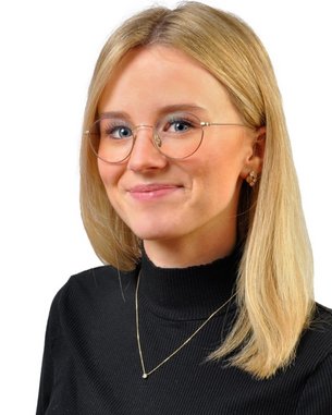 Eine junge Frau mit blonden Haaren und Brille trägt ein schwarzes Oberteil. Sie lächelt leicht und steht vor einem weißen Hintergrund.