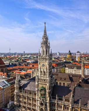 Studienzentrum München | SRH Fernhochschule Moderne Gebäude mit großen Fenstern und einem überdachten Außenbereich. Tische und Stühle unter grünen Sonnenschirmen sind im Vordergrund sichtbar.