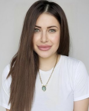 Eine Person mit langen, braunen Haaren und einem weißen T-Shirt lächelt in die Kamera, trägt eine Halskette mit einem Anhänger.