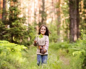 Ein Mädchen mit langen, braunen Haaren trägt einen Pullover mit einem Eulenmotiv und hält einen Zweig in der Hand, während es fröhlich einen Waldweg entlangläuft.