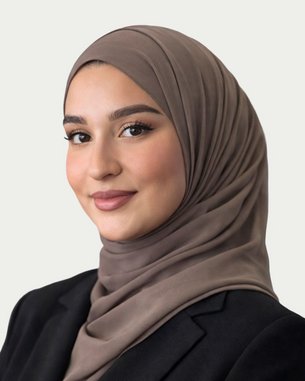 Eine junge Frau mit einem braunen Hijab und einem schwarzen Blazer, die in die Kamera lächelt. Der Hintergrund ist neutral gehalten.