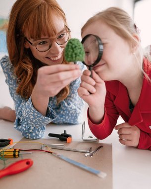 Eine Lehrerin zeigt einem Kind mit einer Lupe eine grüne Bastelarbeit. Auf dem Tisch liegen verschiedene Bastelmaterialien und Werkzeuge. Im Hintergrund ist eine Weltkarte sichtbar.