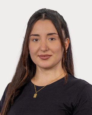 Eine junge Frau mit langen, dunklen Haaren trägt ein schwarzes T-Shirt und eine goldene Halskette. Sie schaut direkt in die Kamera.