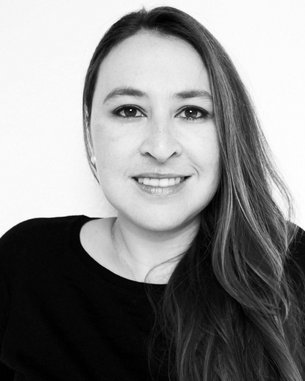 Eine Frau mit langen, braunen Haaren und einem freundlichen Lächeln trägt ein schwarzes Oberteil. Der Hintergrund ist neutral und hell.