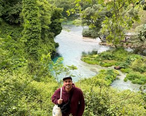 Ein Mann steht lächelnd in einer grünen Landschaft mit Bäumen und einem klaren Fluss im Hintergrund. Er trägt einen roten Hoodie und hält eine Tasche.