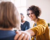 Eine lächelnde Frau mit lockigem Haar trägt einen gelben Pullover und legt ihre Hand auf die Schulter einer anderen Person. Im Hintergrund sind zwei weitere Personen zu sehen, die an einem Gespräch teilnehmen.