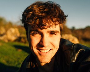 Ein junger Mann mit lockigem Haar lächelt in die Kamera, während er im Freien sitzt. Die Sonne beleuchtet sein Gesicht.