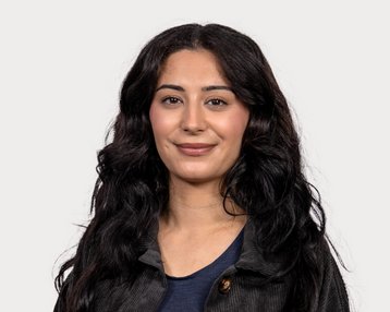 Eine Frau mit langen, lockigen, schwarzen Haaren lächelt in die Kamera. Sie trägt ein schwarzes Oberteil und hat einen neutralen Hintergrund.