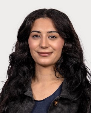 Eine Frau mit langen, lockigen, schwarzen Haaren lächelt in die Kamera. Sie trägt ein schwarzes Oberteil und hat einen neutralen Hintergrund.