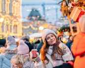Eine Frau mit einer Tasse in der Hand steht in einer belebten Weihnachtsmarkt-Szene, umgeben von festlich beleuchteten Ständen und Menschen.