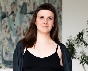 Eine Person mit langen, braunen Haaren trägt ein schwarzes Top und eine schwarze Jacke, steht vor einem abstrakten Kunstwerk und einer Pflanze.