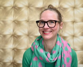 Eine Frau mit Brille und einem bunten Schal mit Herzen lächelt vor einem goldfarbenen, strukturierten Hintergrund.