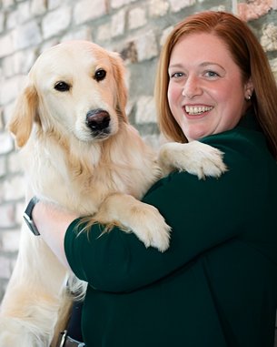 Eine Frau mit langen, roten Haaren hält einen Golden Retriever in ihren Armen. Sie lächelt und steht vor einer gemauerten Wand.