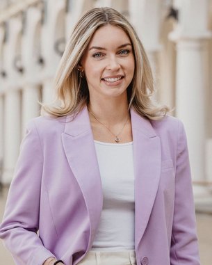 Eine Frau mit langen, blonden Haaren trägt einen lila Blazer und eine weiße Bluse. Sie steht in einer architektonisch gestalteten Umgebung mit Säulen und einem gepflasterten Boden.
