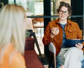 Berufe in der klinischen Psychologie ohne Approbation Eine Frau mit Brille und braunem Blazer sitzt auf einem Stuhl und spricht mit einer anderen Person, während sie ein Notizblatt hält.