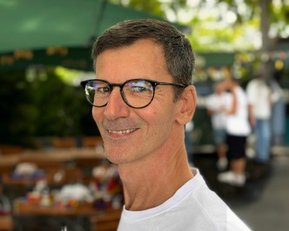 Ein Mann mit Brille und kurzem Haar lächelt in die Kamera. Er trägt ein weißes T-Shirt und steht vor einem Markt mit verschwommenen Personen im Hintergrund.